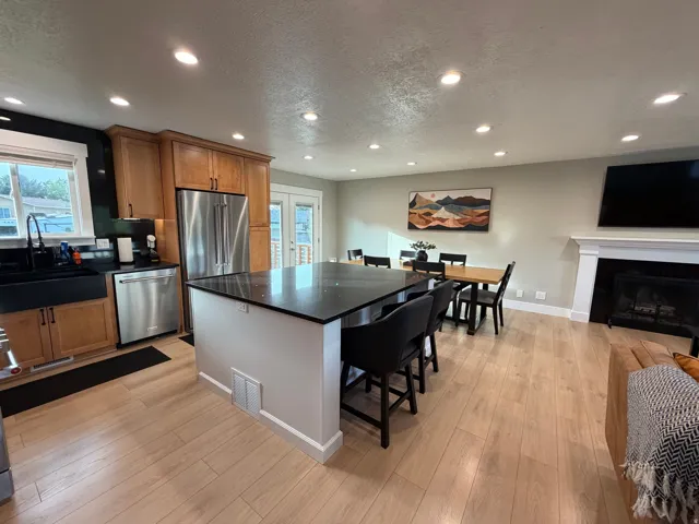 Open-concept living area featuring wood-finish flooring, a kitchen island with dark countertops, stainless steel appliances, and a fireplace with a white mantel