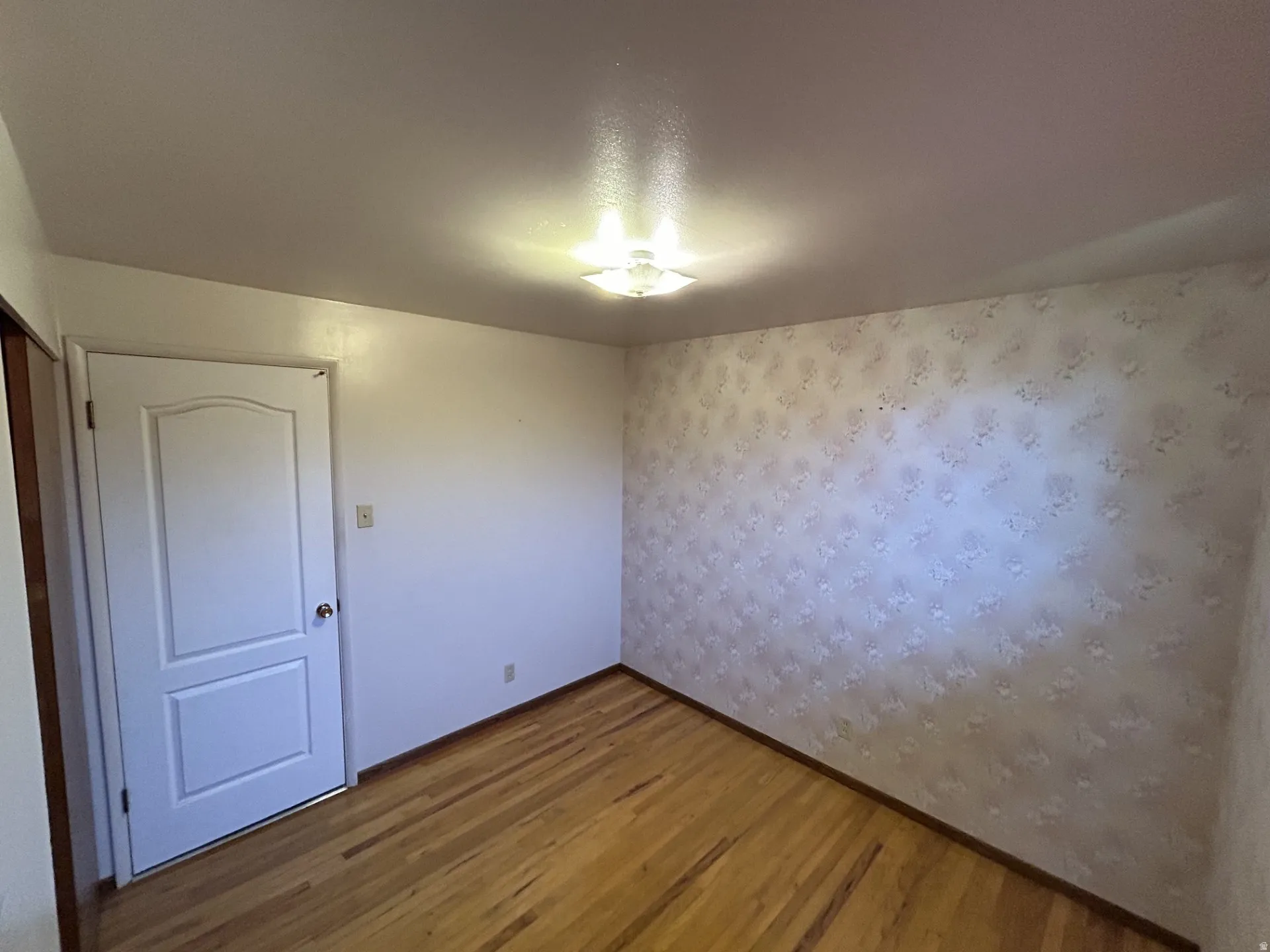 2nd bedroom featuring wood-finish flooring, a white paneled door & patterned wallpaper.