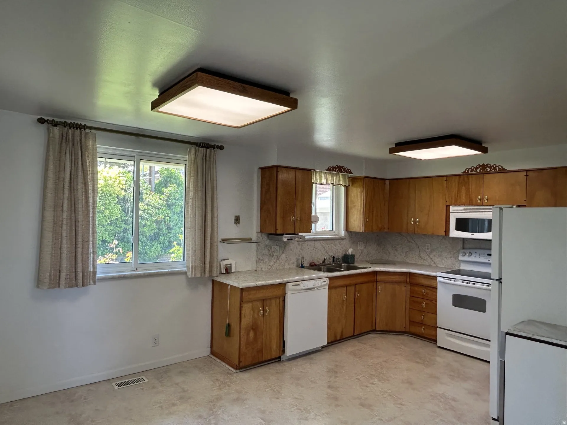 Kitchen featuring 2 light fixtures, wood cabinetry, white appliances, & a double sink.