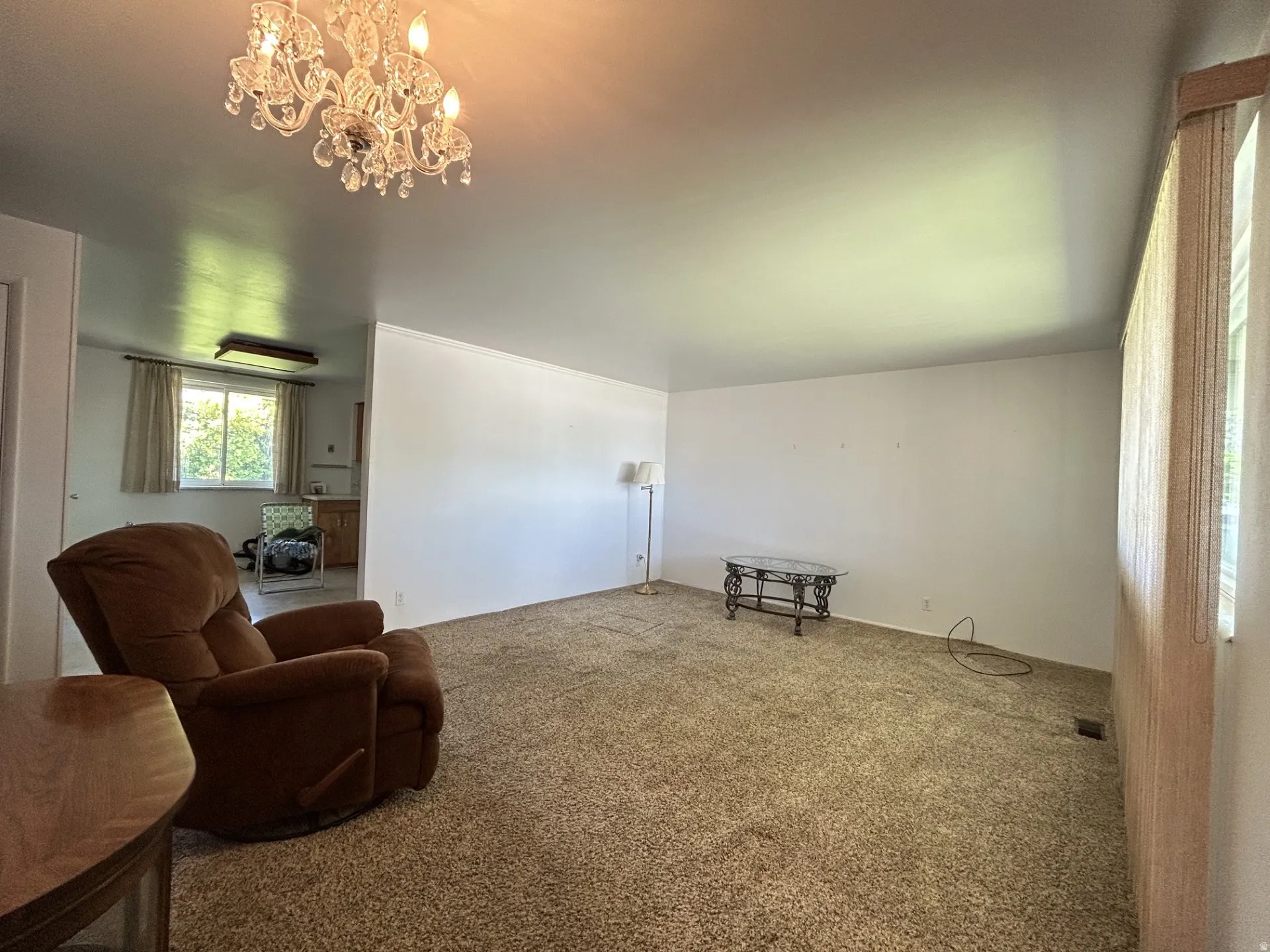 Spacious living area featuring neutral tone carpeting, a decorative chandelier & abundant natural light from the large window.