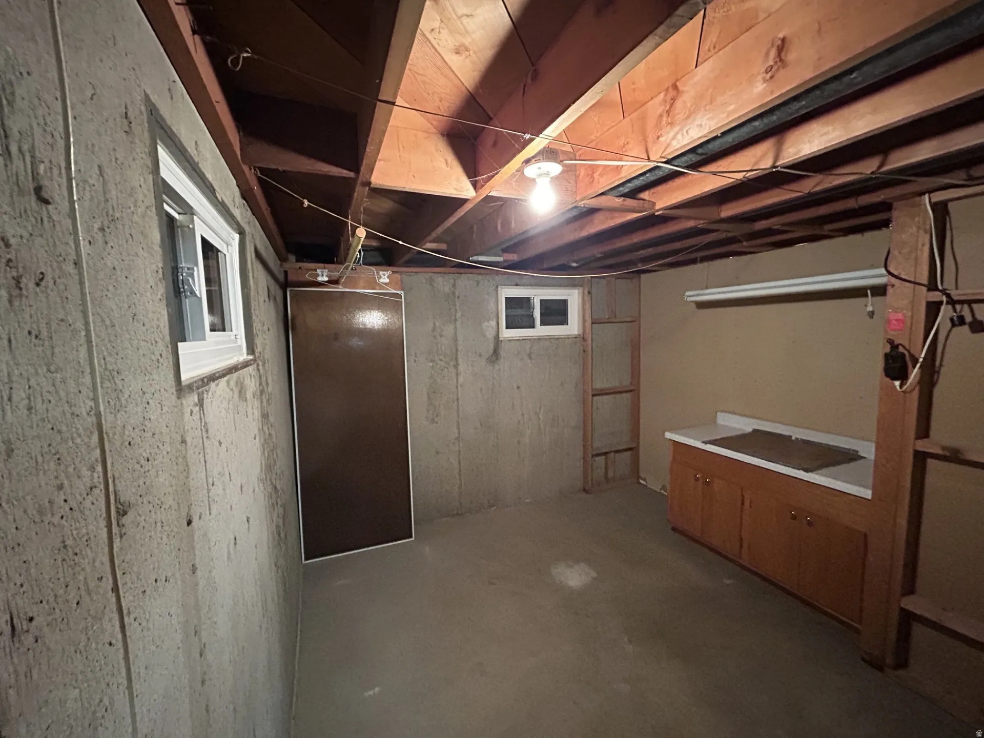Basement utility area featuring concrete walls, exposed ceiling joists, two windows with white frames, a utility sink with wood cabinetry, and a built-in shelving unit