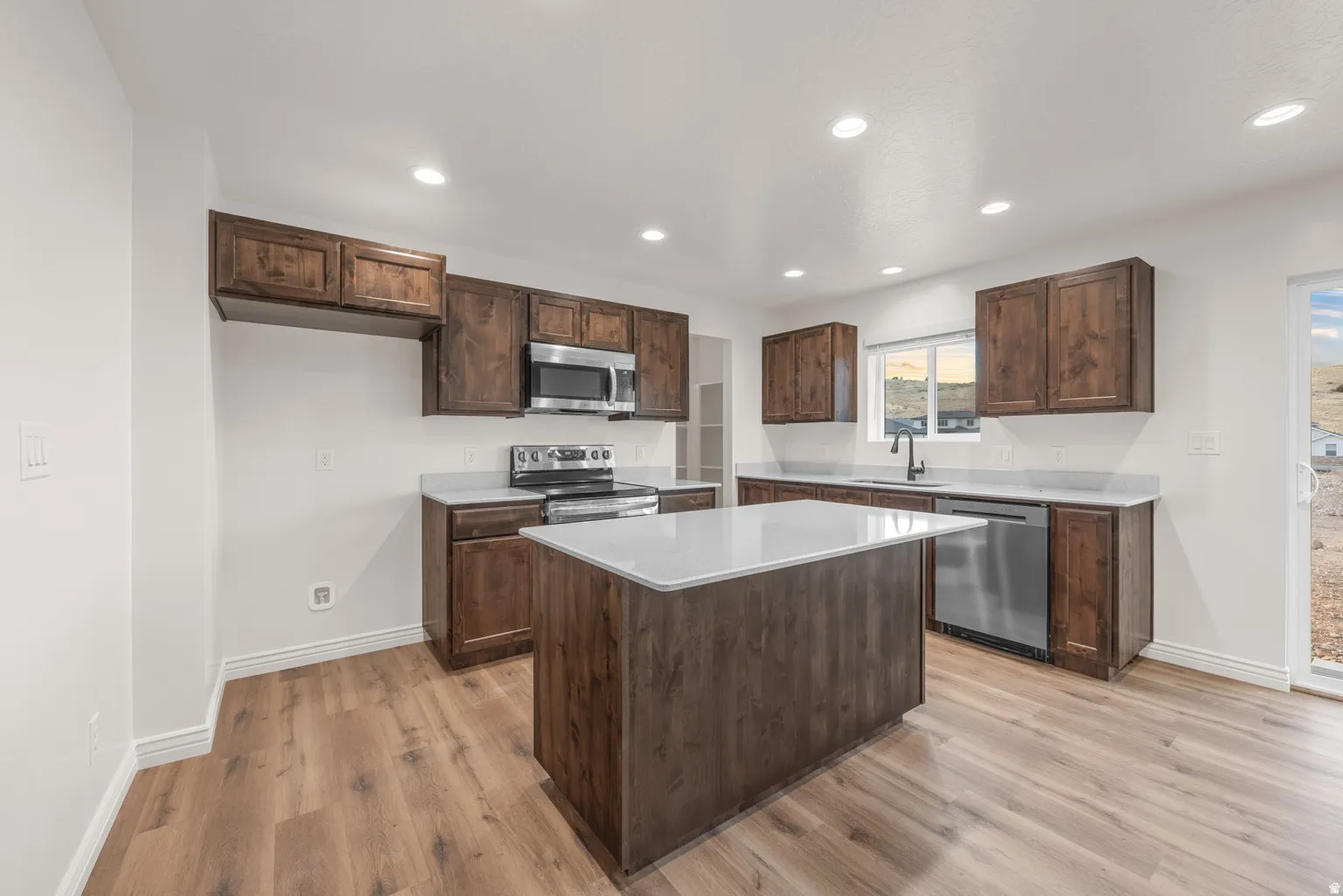 Modern kitchen featuring wood-finish flooring, white walls, dark wood cabinetry, stainless steel appliances, and light-toned countertops