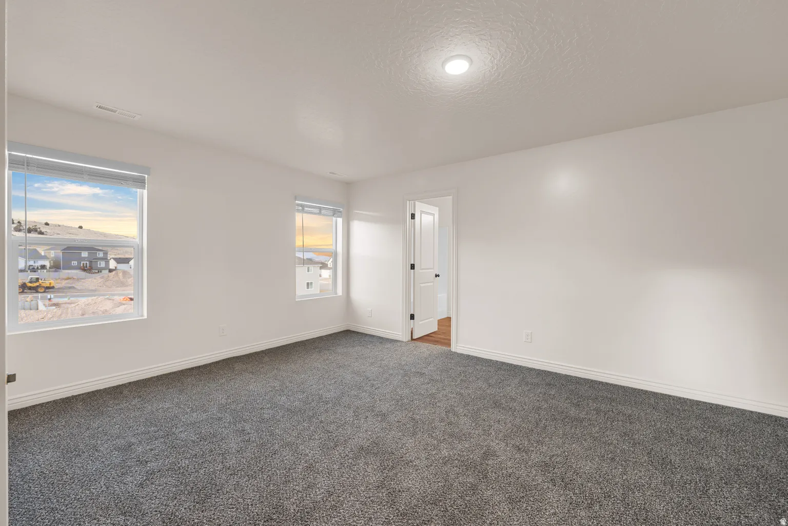 Spacious room featuring neutral paint, gray carpeting, dual windows with roller blinds, a recessed ceiling light fixture, and white baseboards