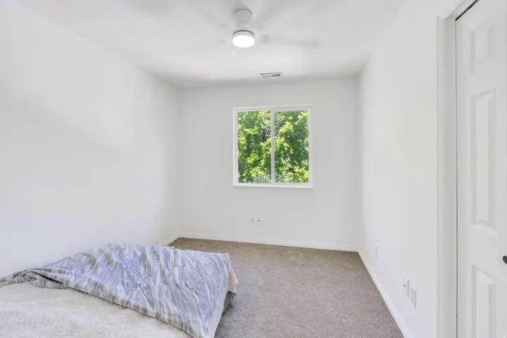Bright room featuring a window overlooking greenery, neutral carpet flooring, white walls, and a ceiling fan with integrated lighting