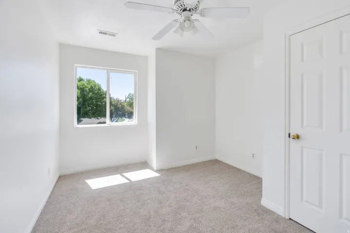 Bright room featuring a large window, neutral carpeting, a ceiling fan, white paneled door, and white baseboards