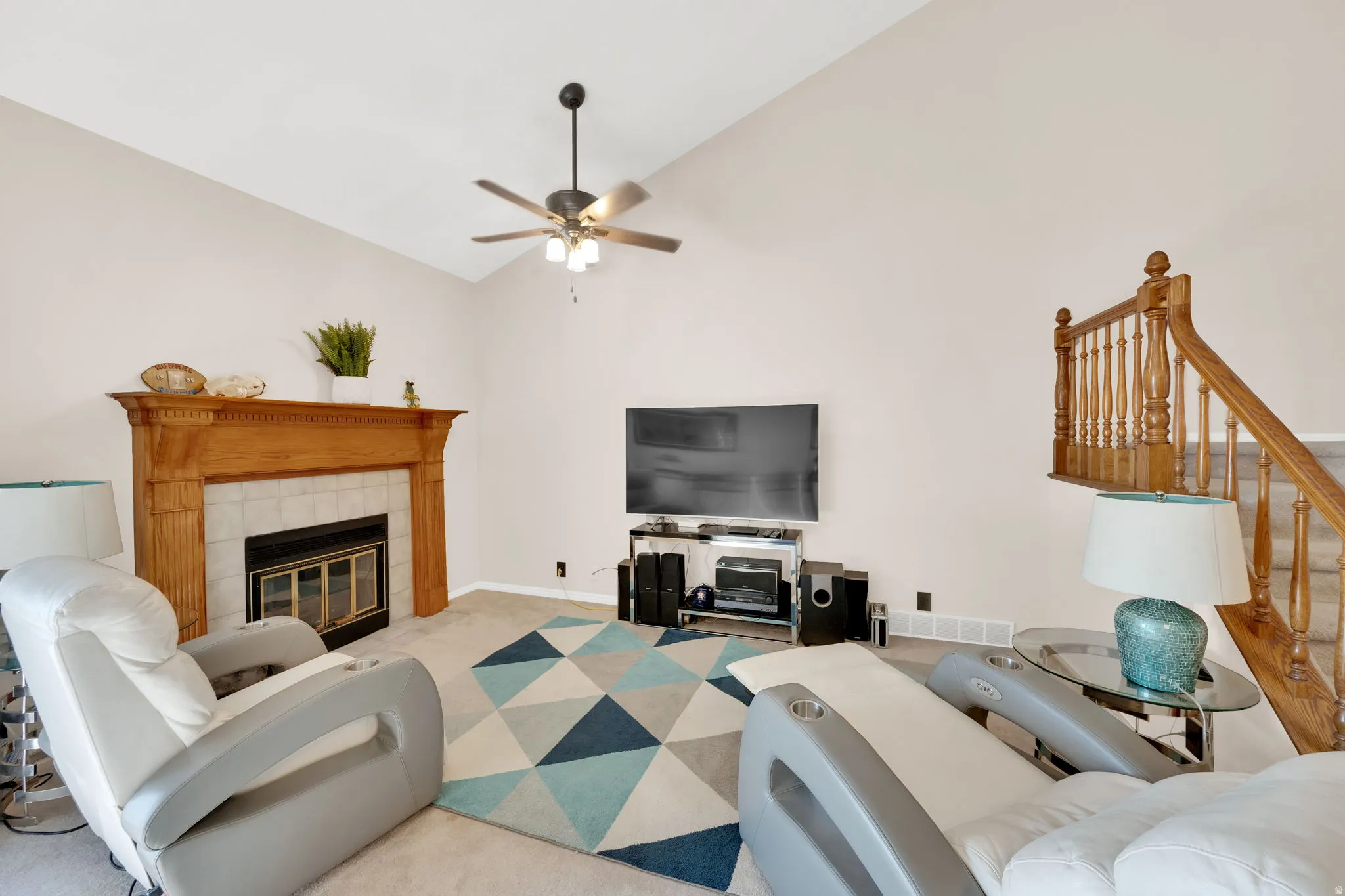 Living area featuring a vaulted ceiling with a ceiling fan, a fireplace with a wood-finish mantel and tiled surround, and a wooden staircase with baluster railing