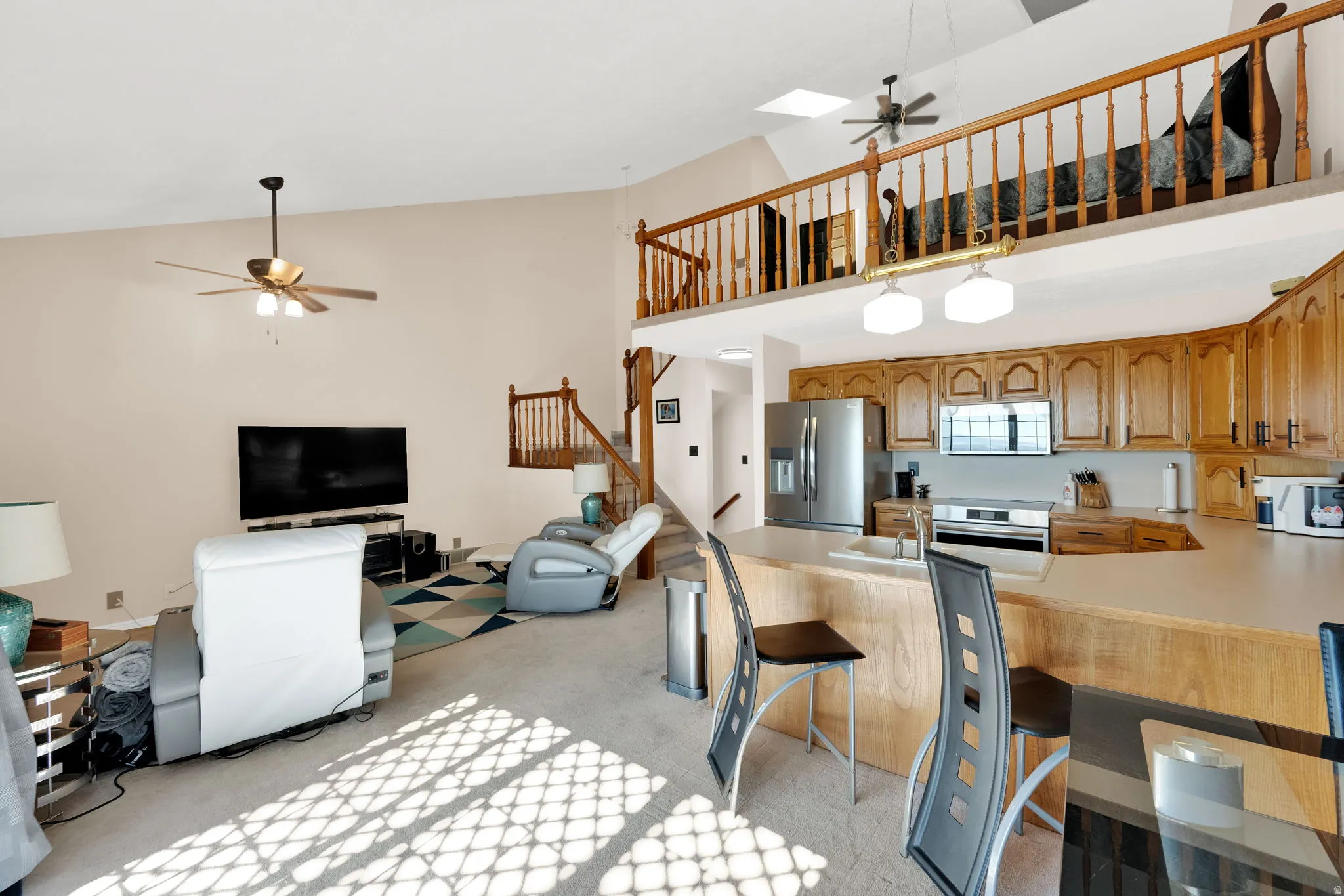 Open-concept living area featuring a vaulted ceiling with skylights, an upper-level loft with wood railing, a kitchen with wood cabinetry and stainless steel appliances, and a breakfast bar