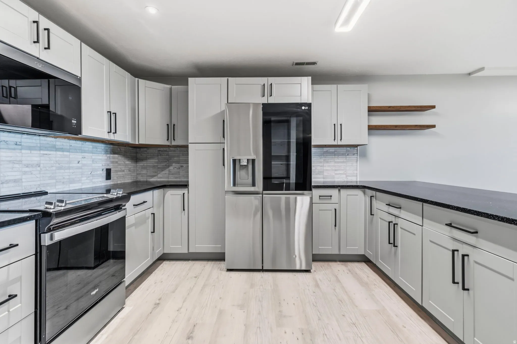 Modern kitchen featuring light gray cabinetry, stainless steel appliances, black countertops, a tile backsplash, and light wood-finish flooring
