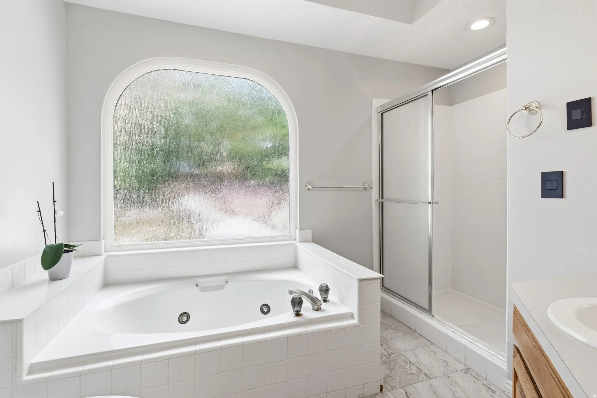 Soaking tub with jetted features, an arched frosted window, a glass-enclosed shower, a wood-finish vanity, and recessed lighting