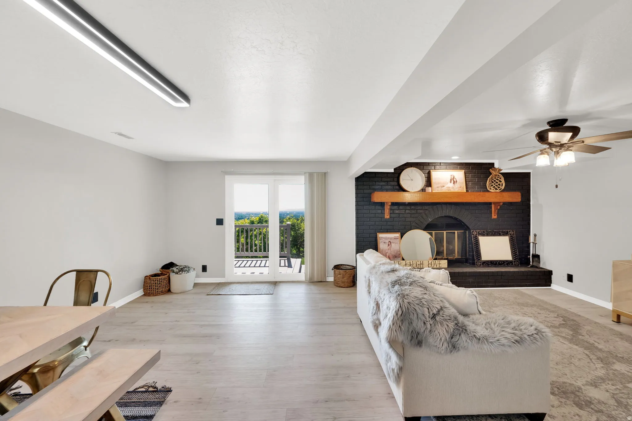 Spacious living area featuring light wood-finish flooring, a black brick fireplace with a wood mantel, and direct access to an outdoor deck through sliding glass doors