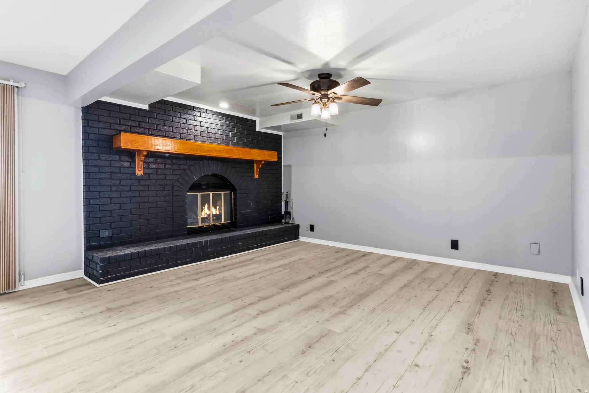 Expansive room featuring wood-finish flooring, a black brick fireplace with an arched firebox, a wood mantel, and a ceiling fan with integrated lighting