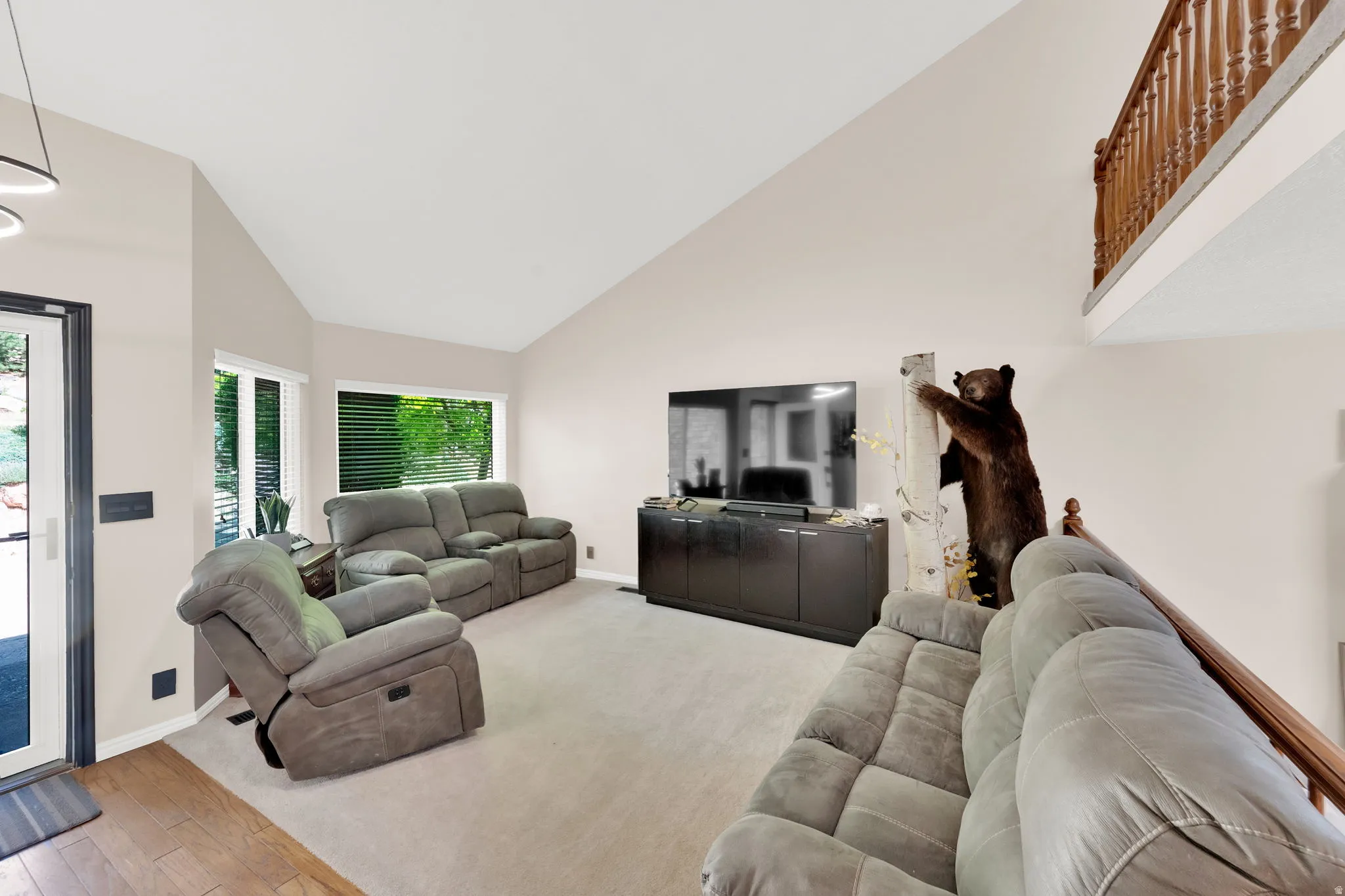 Vaulted ceiling living room featuring a wood-finish entry, carpeted flooring, and multiple windows with horizontal blinds