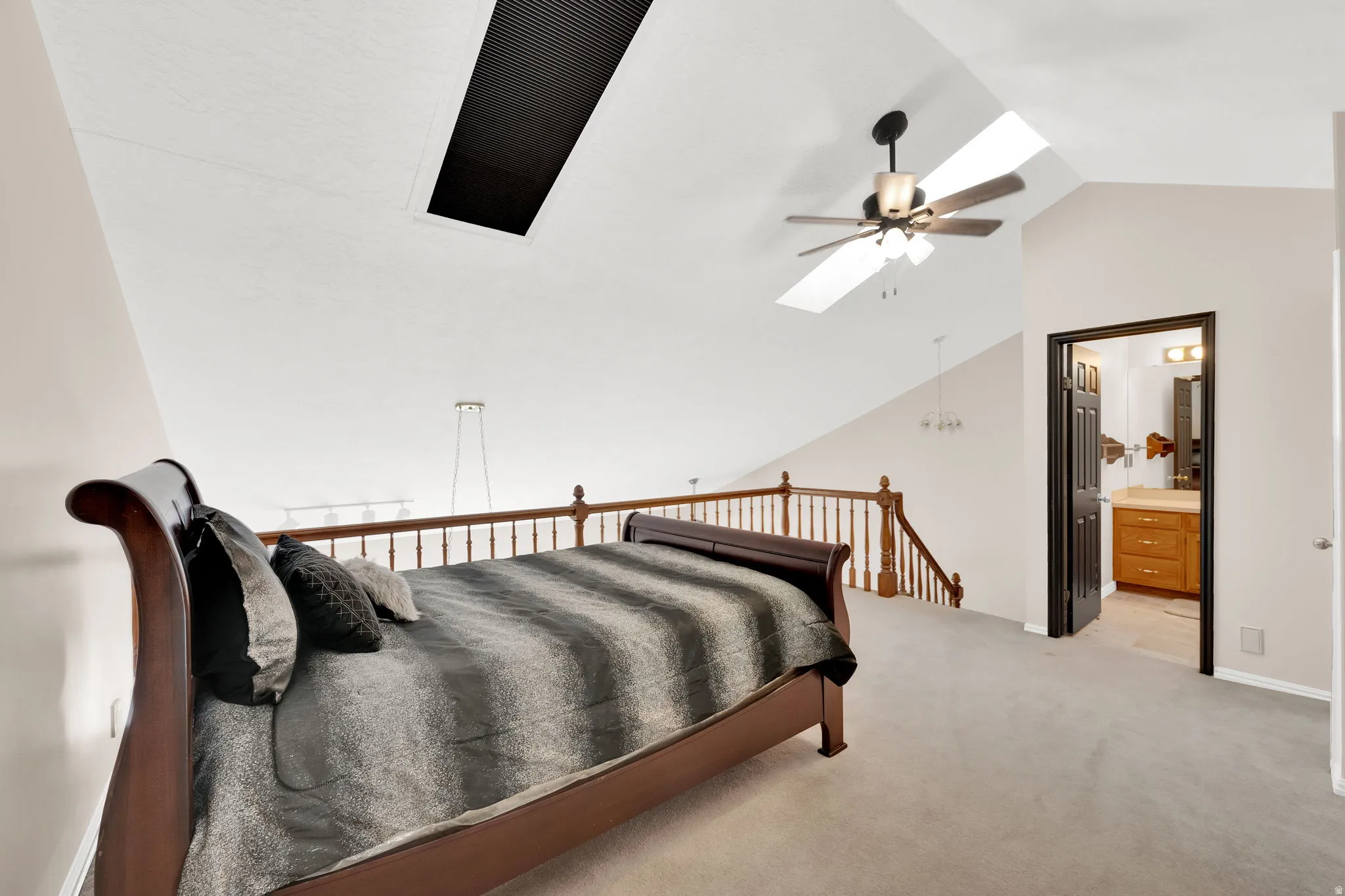 Vaulted ceiling with skylight and ceiling fan, featuring light gray carpet, wood spindle railing, and an adjacent bathroom with a wood vanity and mirror