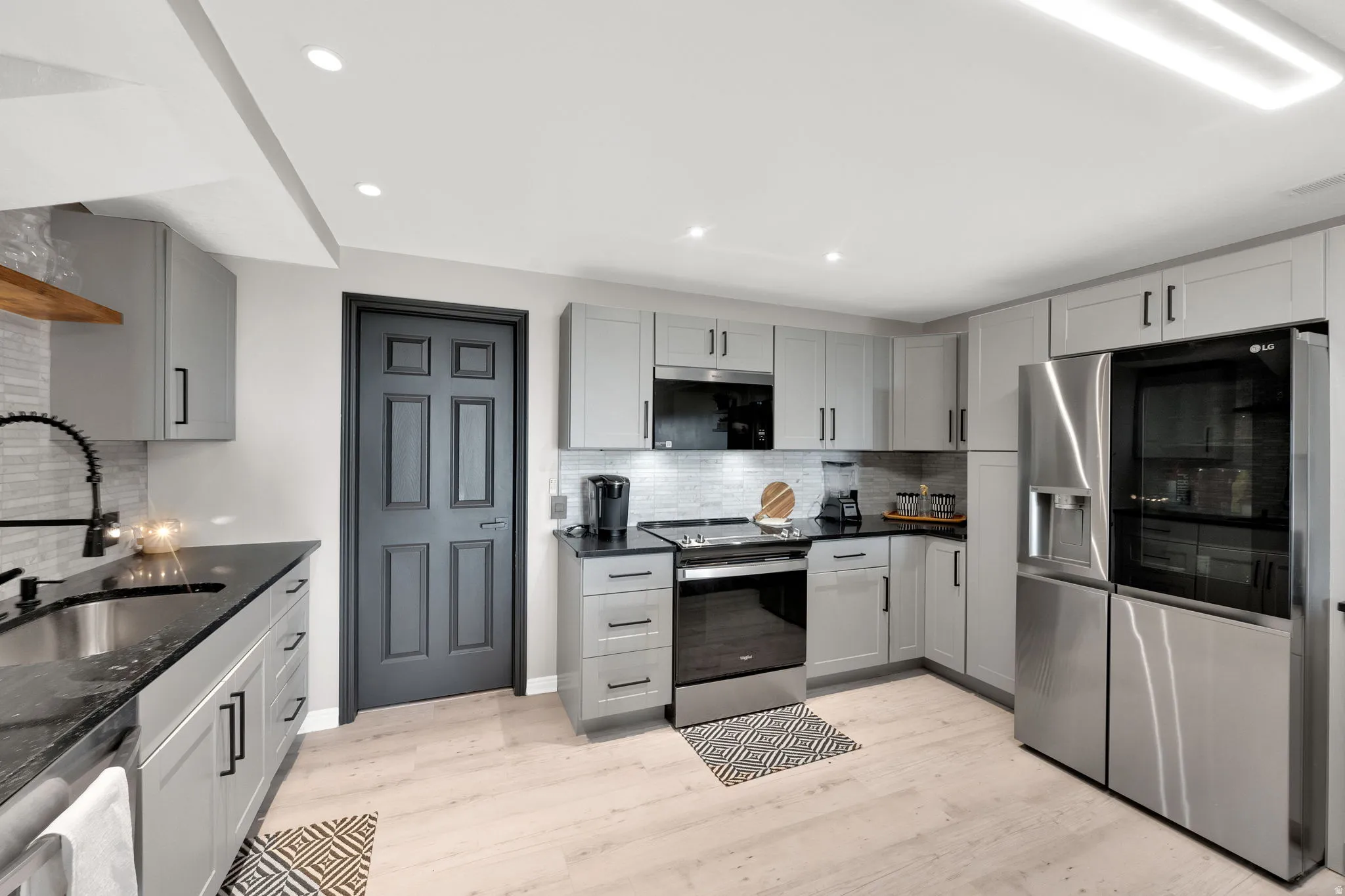 Modern kitchen featuring light wood-finish flooring, grey cabinetry with black hardware, black countertops, and stainless steel appliances