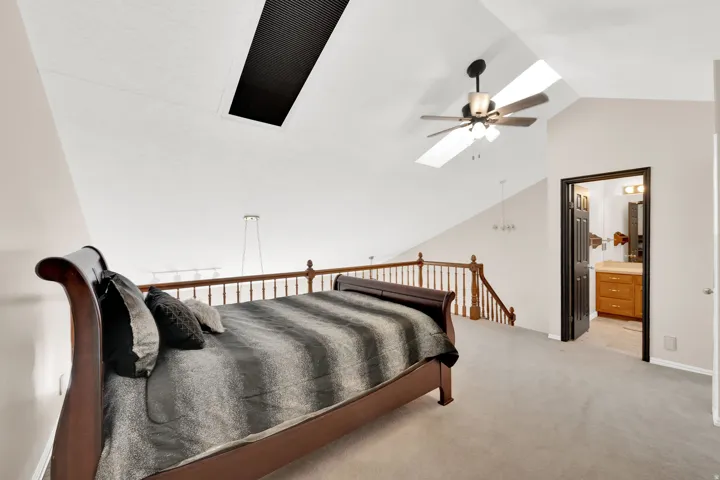 Vaulted ceiling with skylight and ceiling fan, featuring light gray carpet, wood spindle railing, and an adjacent bathroom with a wood vanity and mirror