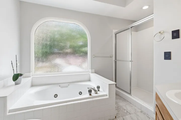 Soaking tub with jetted features, an arched frosted window, a glass-enclosed shower, a wood-finish vanity, and recessed lighting