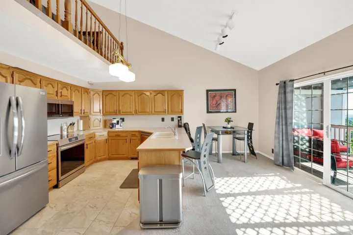 Open-concept kitchen and dining area featuring light wood cabinetry, stainless steel appliances, tile flooring, a vaulted ceiling, and direct access to an outdoor deck through sliding glass doors