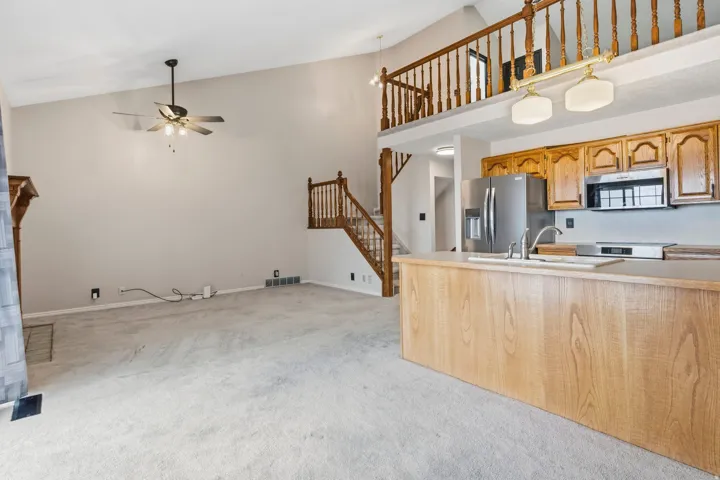 Vaulted ceiling with ceiling fan, wood-finish kitchen cabinetry, stainless steel kitchen appliances, kitchen island with integrated sink, and interior balcony with wood railing