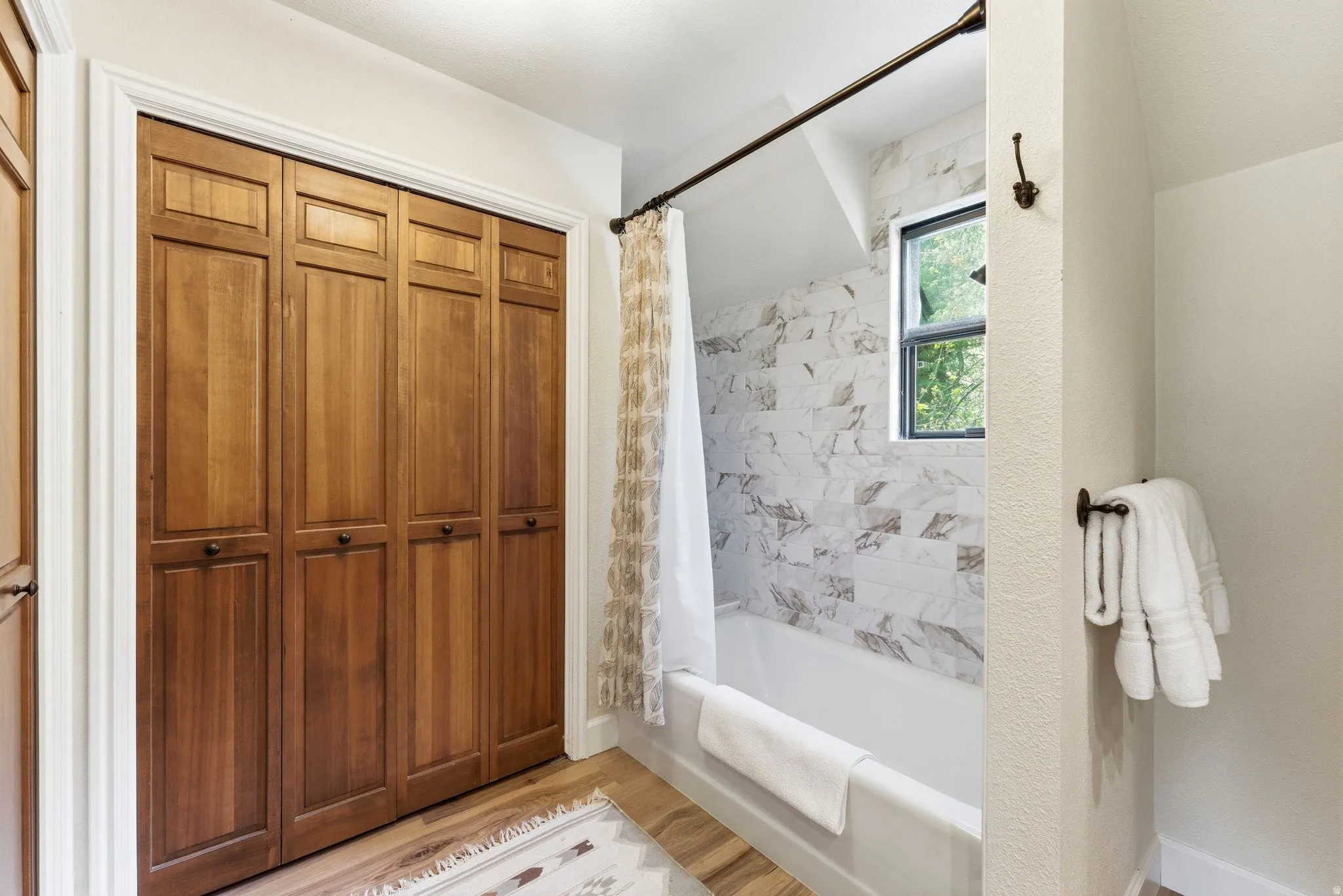 Bathroom featuring a bathtub with marble-look tile surround, a window, wood-finish flooring, and bi-fold closet doors