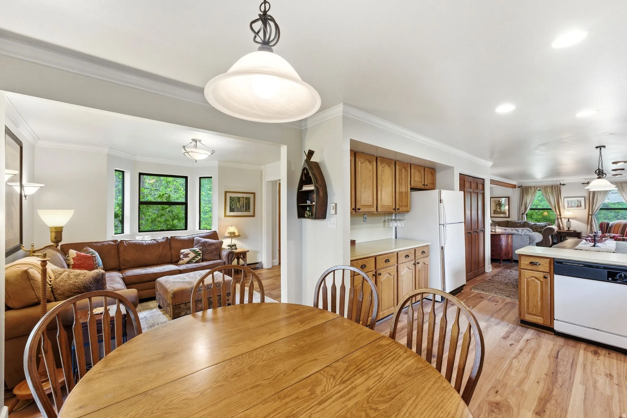 Open-concept living space featuring wood-finish flooring throughout, a dining area with a natural wood table, a kitchen with light wood cabinetry and white appliances, a breakfast bar, and a living room with a bay window