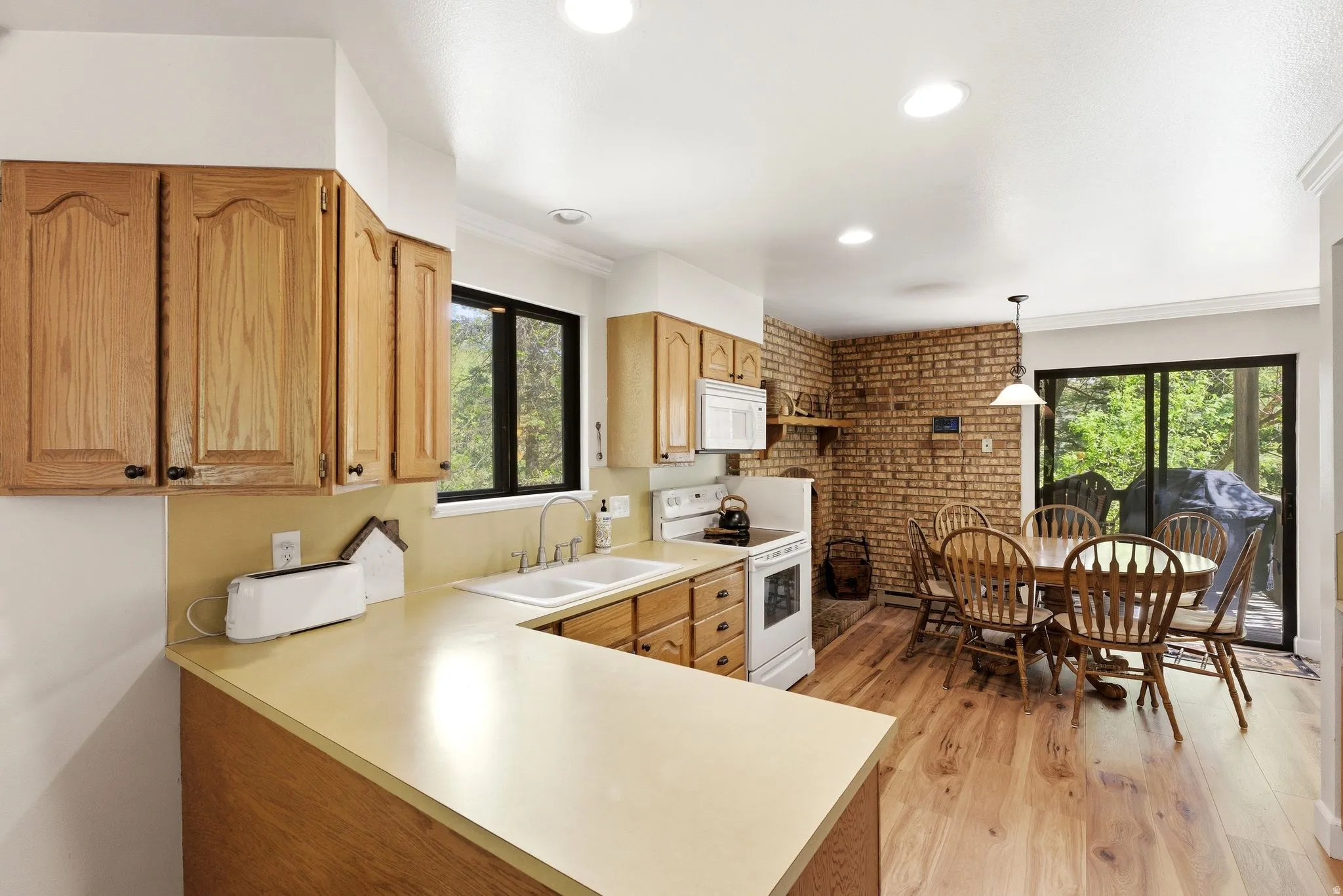 Open-concept kitchen and dining area featuring wood-finish flooring, brick accent wall, recessed lighting, and sliding glass doors