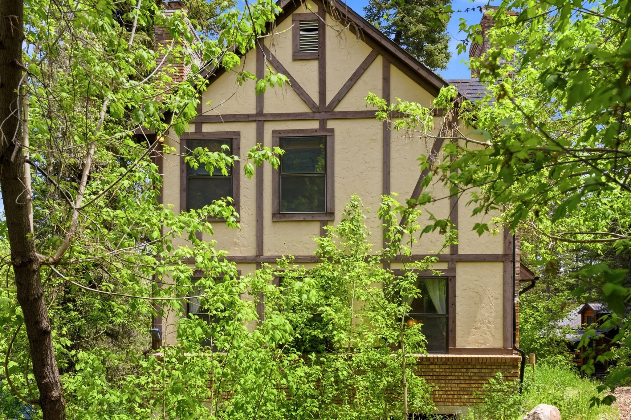 Tudor-style exterior featuring cream stucco with dark wood trim, multiple windows, and a visible portion of a brick foundation