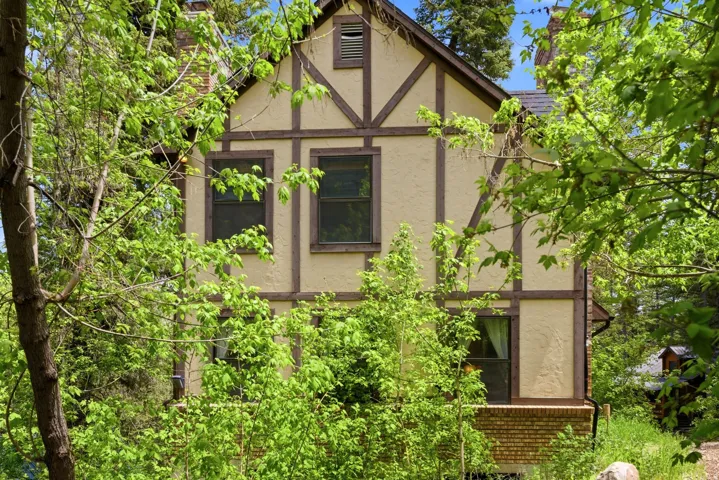 Tudor-style exterior featuring cream stucco with dark wood trim, multiple windows, and a visible portion of a brick foundation