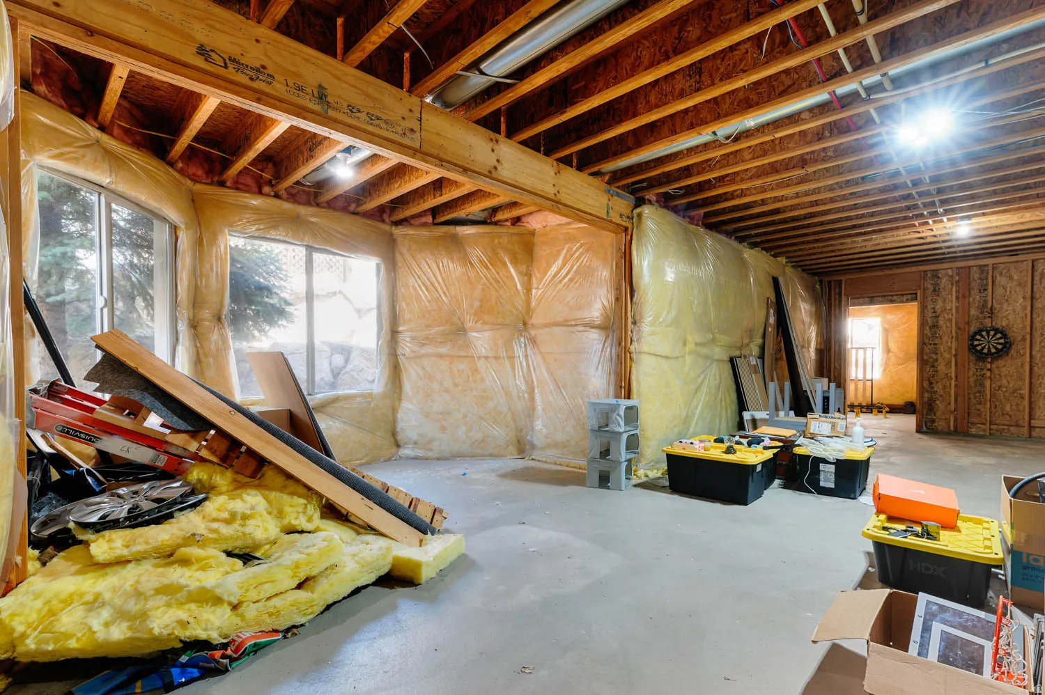 Unfinished basement presenting exposed wood ceiling joists and support beams, multiple windows, and a concrete floor