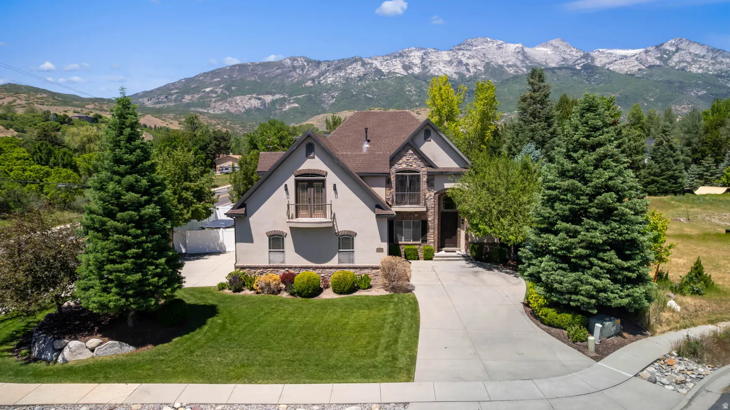 Stucco and stone exterior home featuring a curved driveway, established landscaping, and a mountainous backdrop