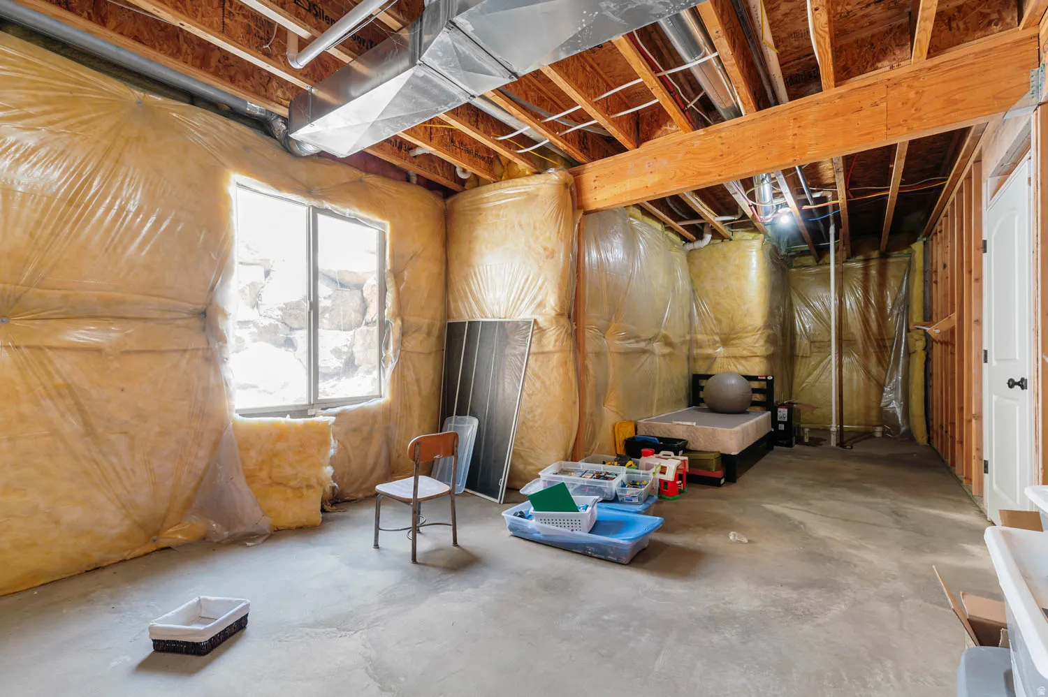Unfinished basement space featuring exposed wooden ceiling joists, concrete flooring, insulated walls, and a window for natural light
