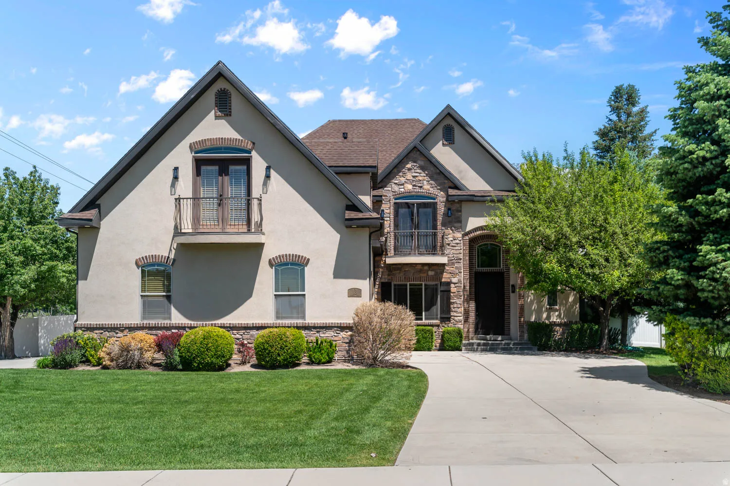 Stucco and stone facade home featuring a prominent arched entry, two upper balconies with wrought iron railings, an expansive front lawn, and a concrete driveway