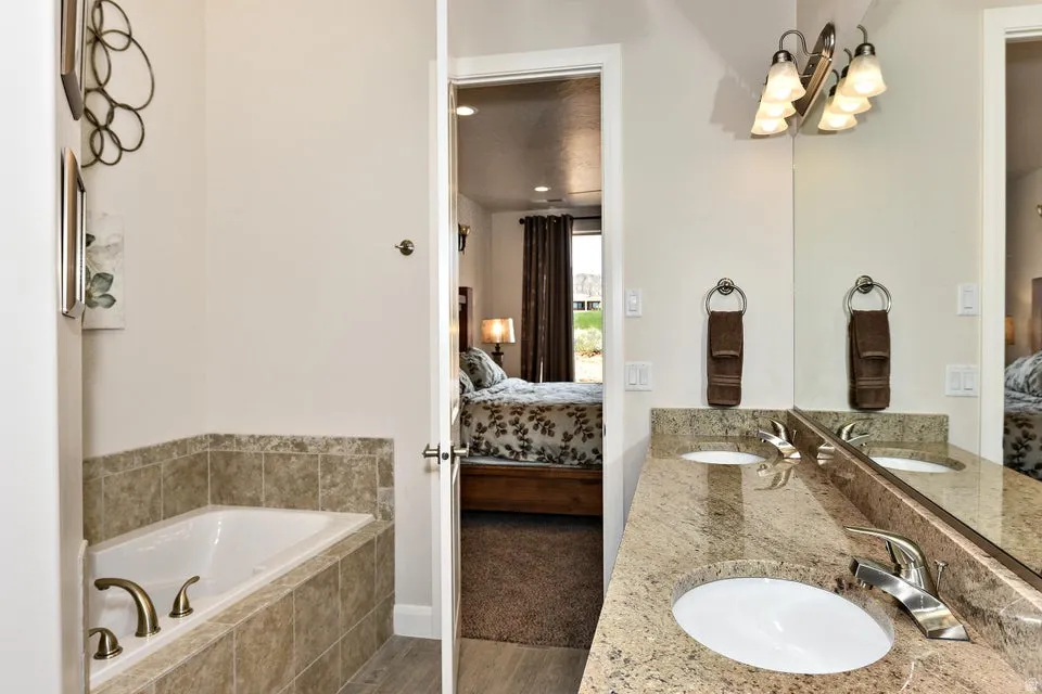 Bathroom featuring a built-in soaking tub with brass fixtures, a dual vanity with light-toned stone countertop, integrated sinks, and a large mirror