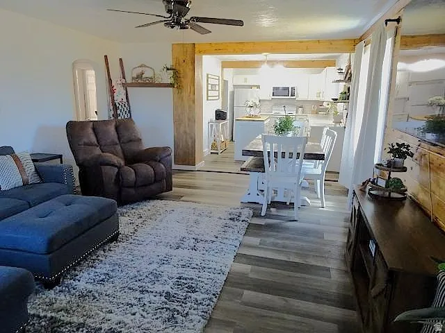 Open-concept living space featuring wood-finish flooring, a ceiling fan with integrated lighting, and a wide entryway with exposed wood beams