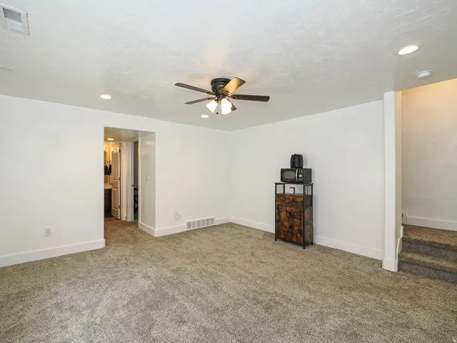 Carpeted room featuring a ceiling fan with integrated lighting, recessed ceiling lights, a doorway to a bathroom, and a carpeted staircase