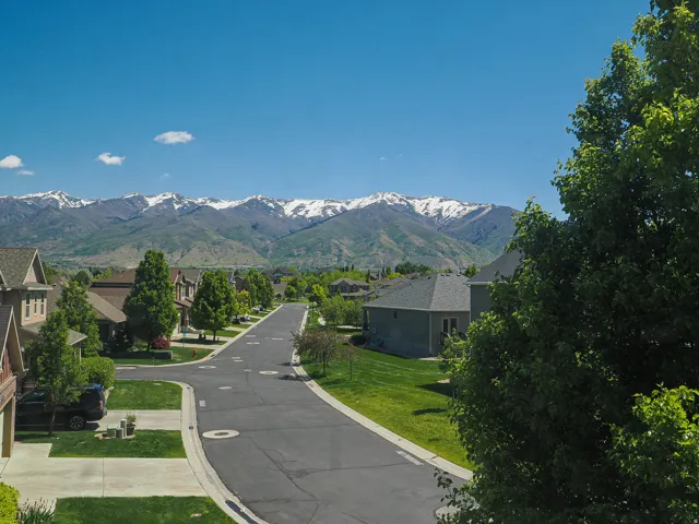 Residential street with manicured lawns and mature trees, featuring distant snow-capped mountains and a clear sky