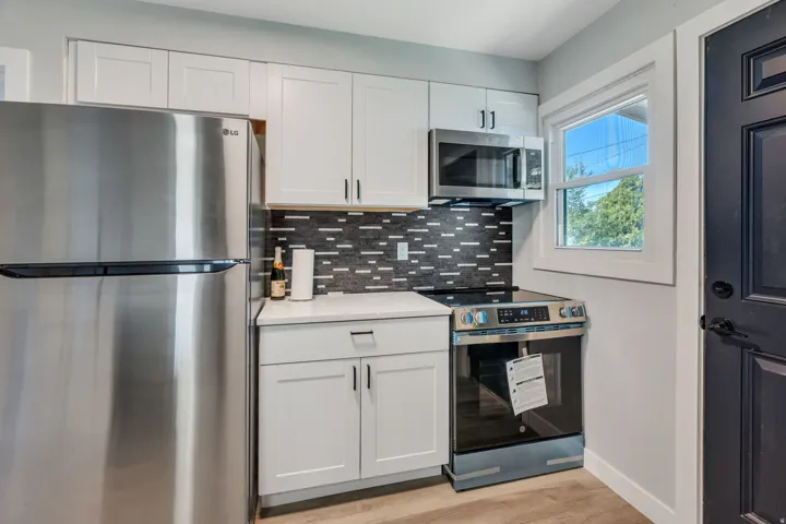 Kitchen featuring white shaker cabinetry, stainless steel appliances, a mosaic tile backsplash, a window with white trim, and wood-finish flooring