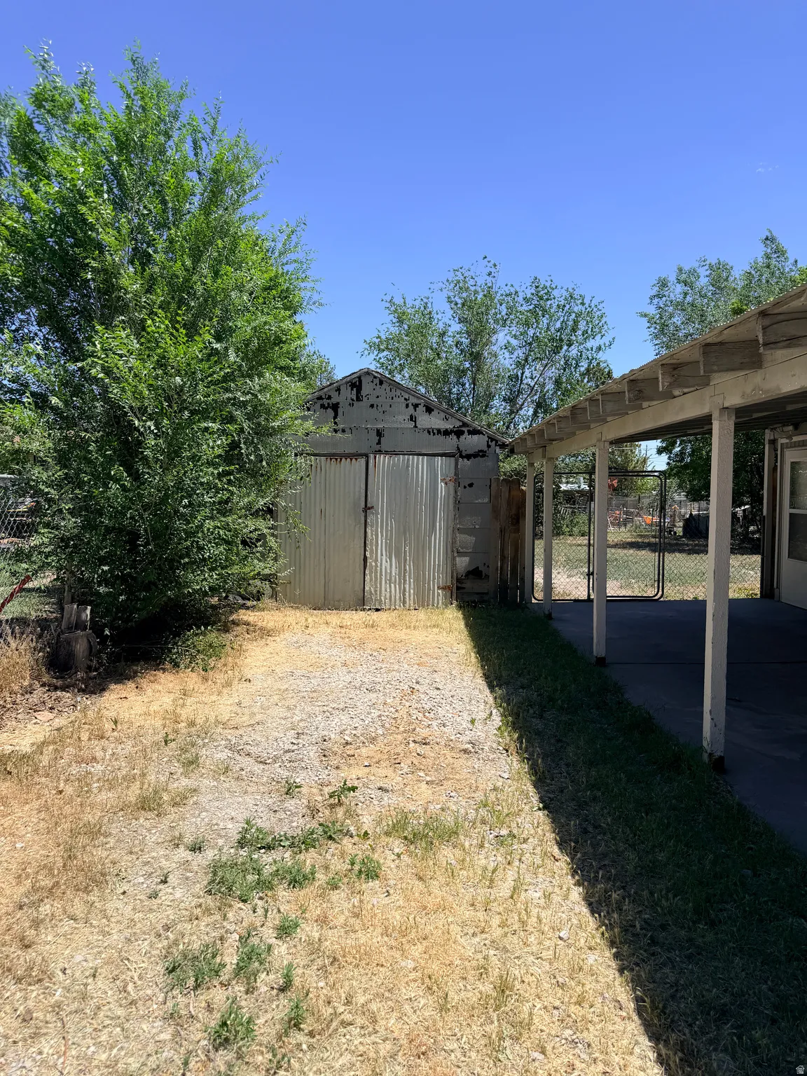 Detached corrugated metal shed with double doors, gravel pathway, and adjacent covered patio featuring exposed rafters and supporting columns