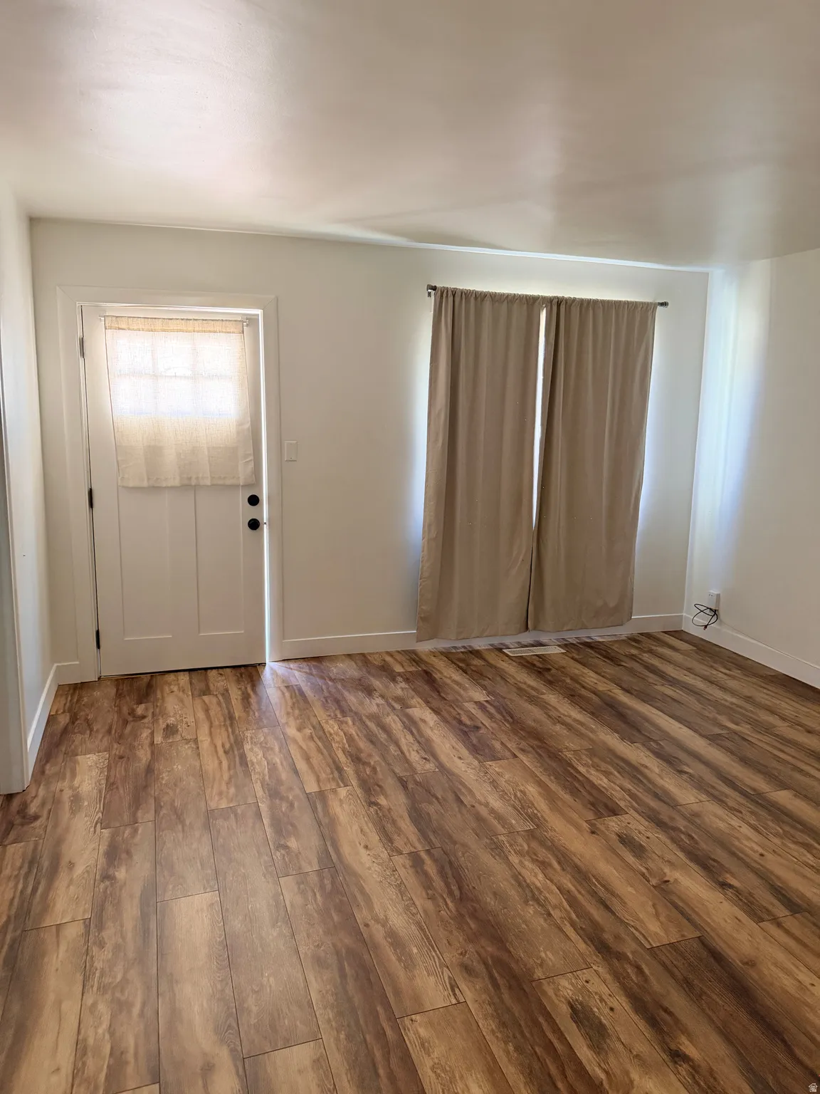 Spacious room featuring wood-finish flooring, white walls, and a white door with black hardware