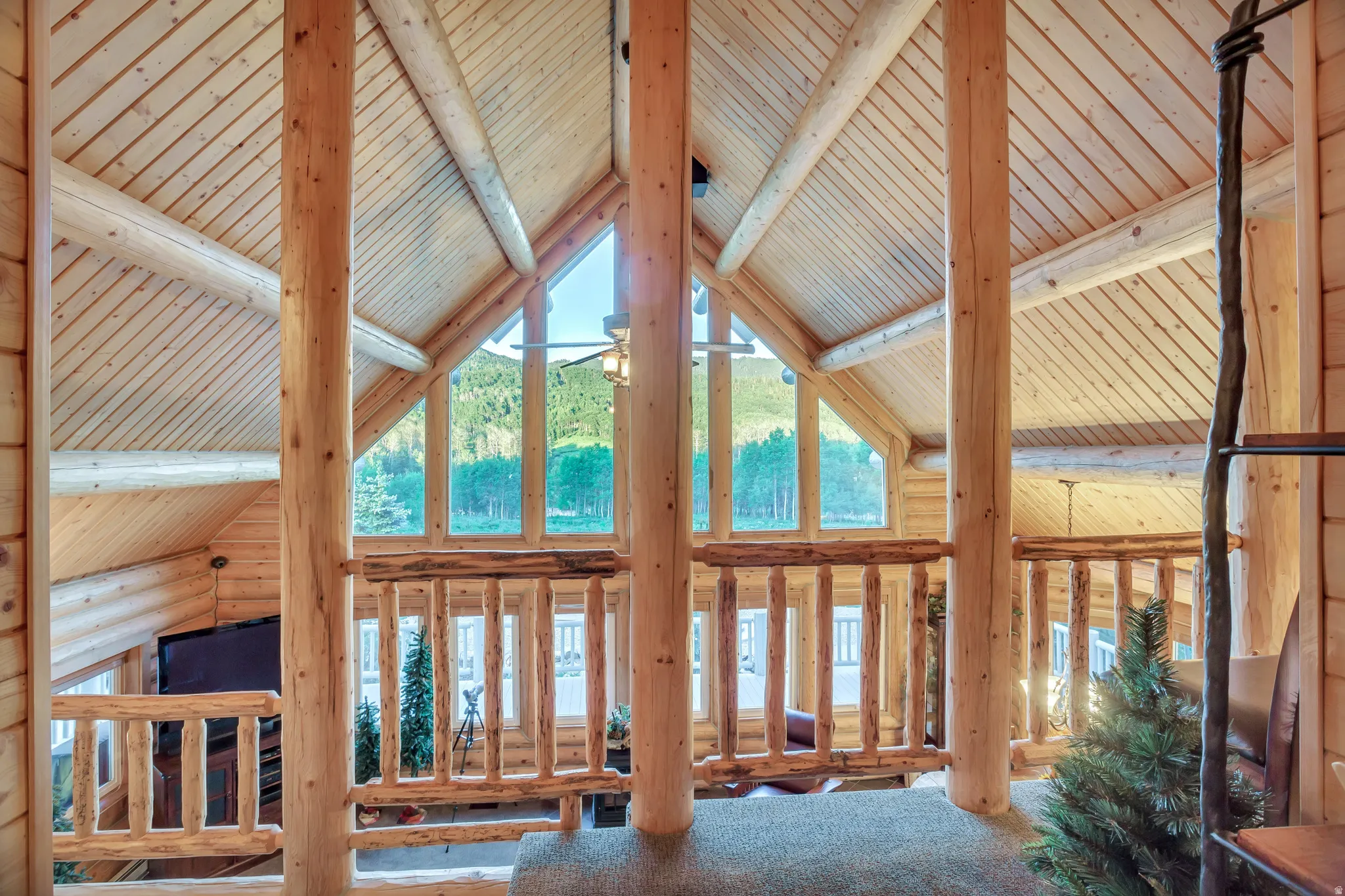 Vaulted ceiling with natural wood planking, exposed log beams, full-height window wall, log railing, and carpeted flooring