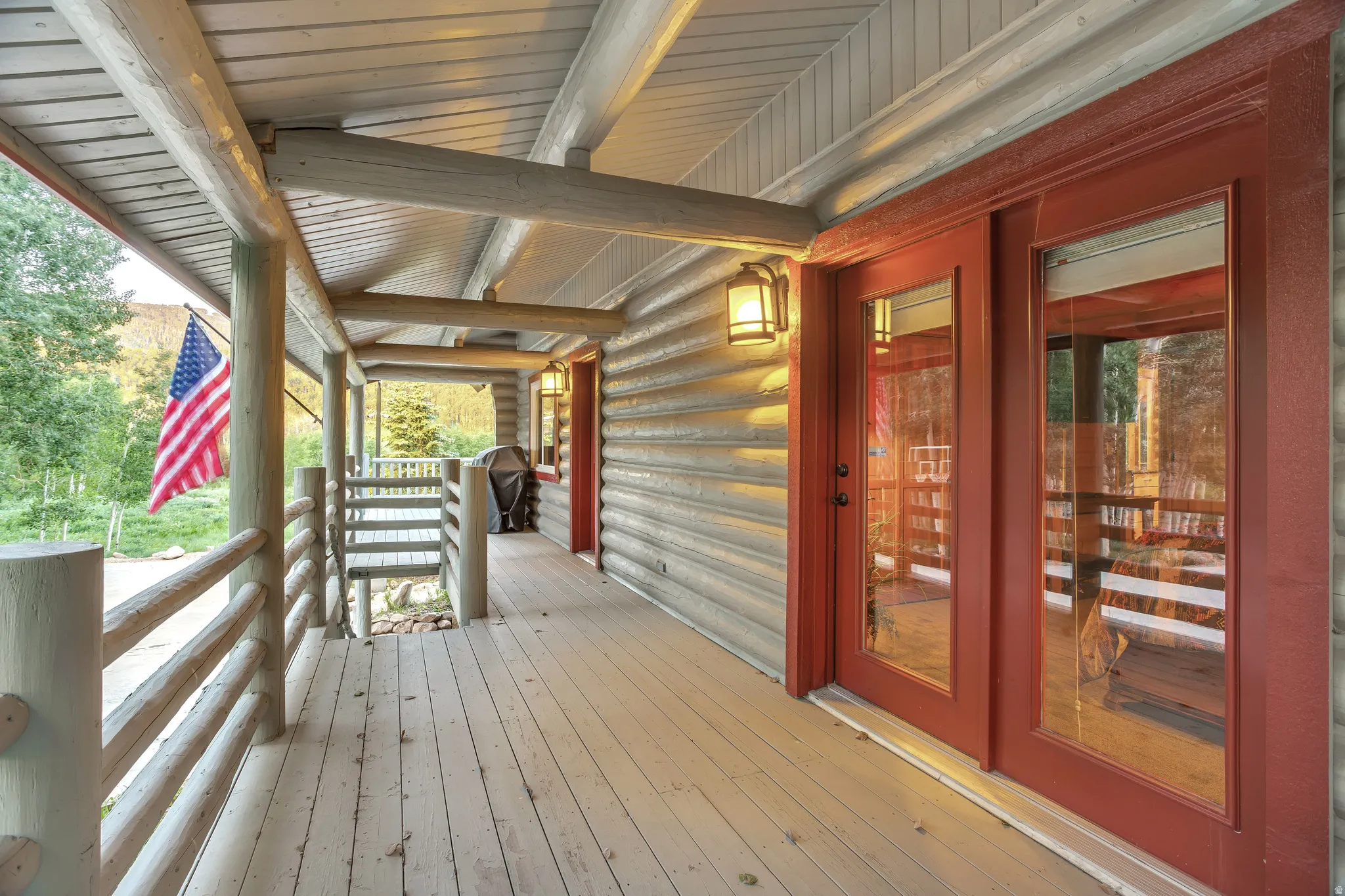 Expansive covered porch featuring a log railing system, wood-finish flooring, log beam ceiling, and a prominent red-framed glass door entry