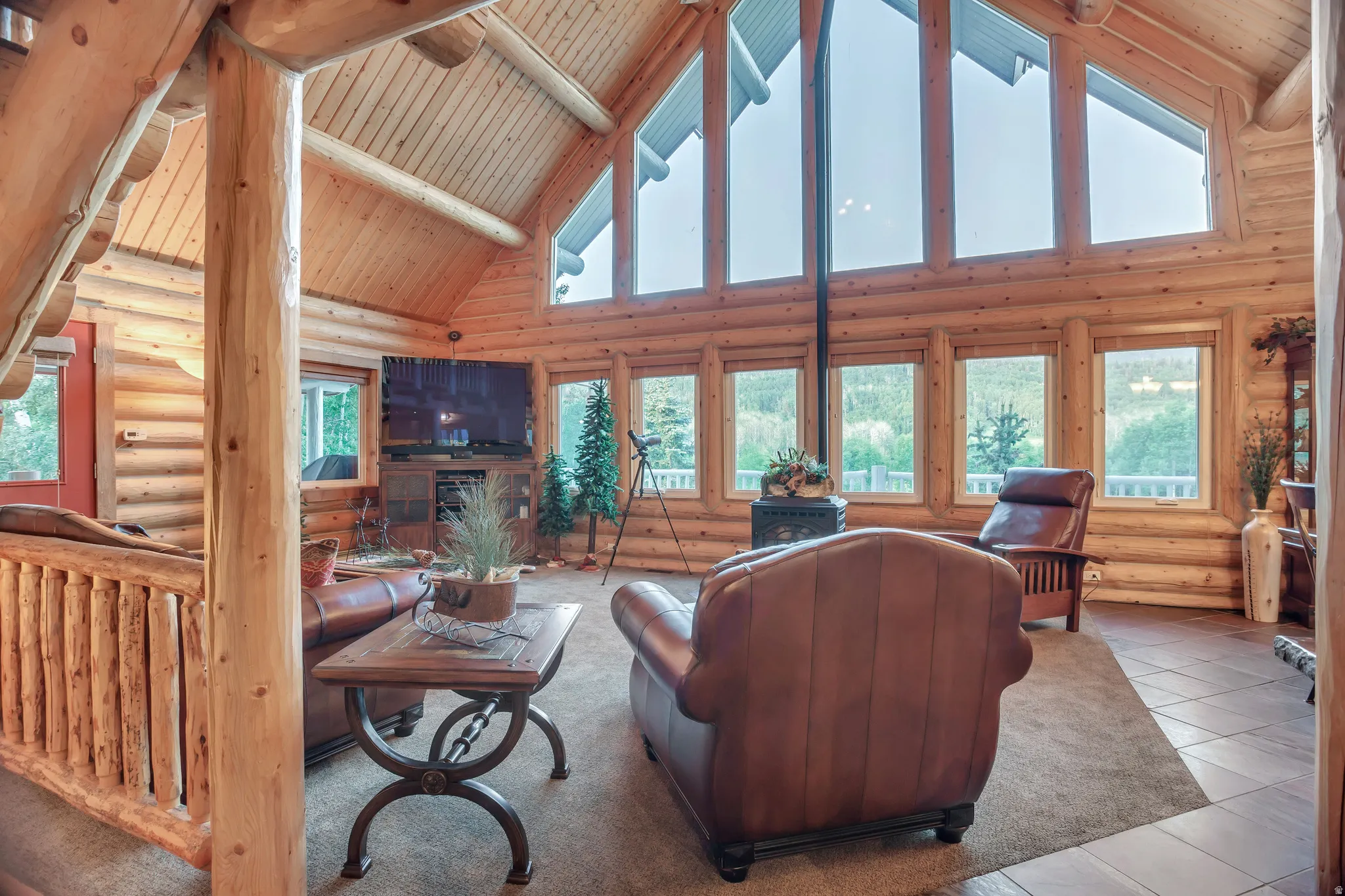 Vaulted wood-finish ceiling with exposed log beams, extensive window array, log construction walls, and a freestanding wood-burning stove