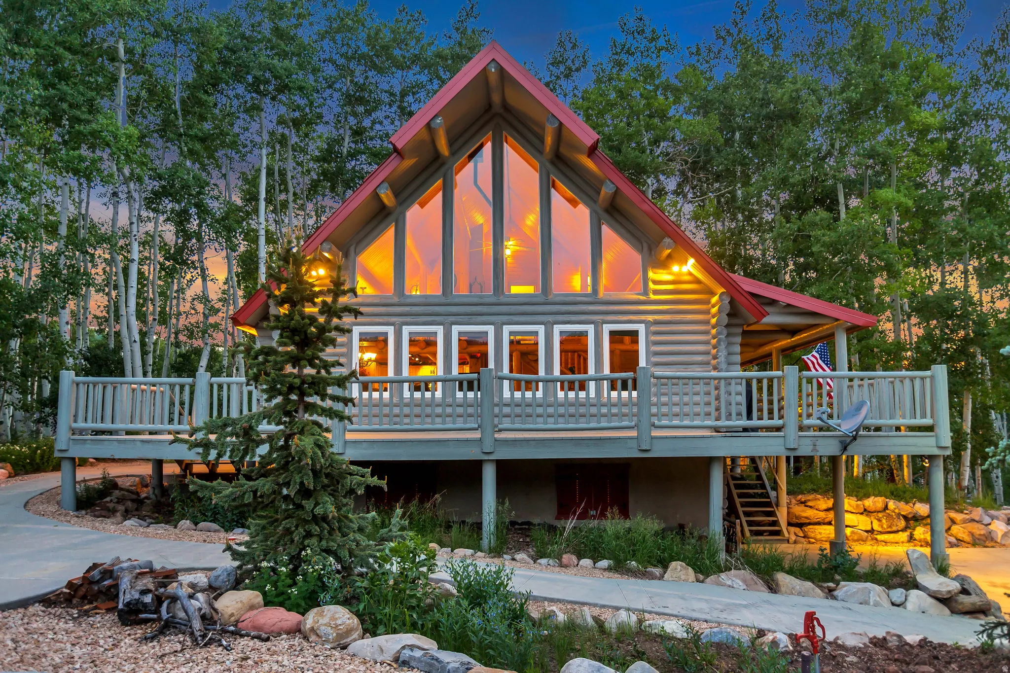 Log cabin style residence featuring a prominent A-frame design, expansive front deck, large picture windows, a red metal roof, and a surrounding natural landscape with mature trees