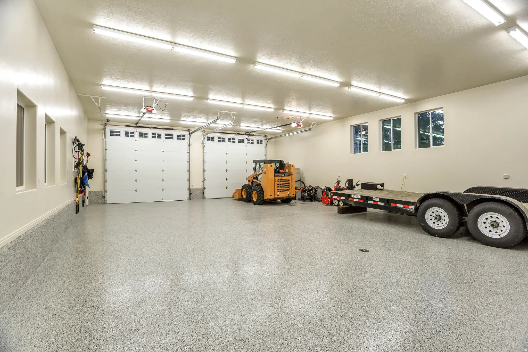 Spacious multi-car garage featuring a speckled epoxy floor, two overhead garage doors, ample wall-mounted shelving, and multiple windows providing natural light