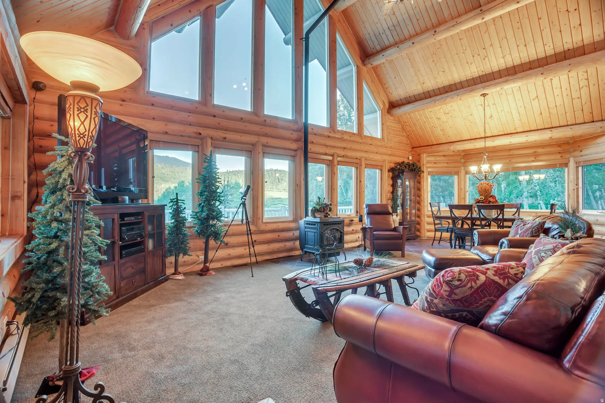 Open-concept living area featuring a vaulted wood-finish ceiling, exposed log beam construction, extensive window array, and carpeted flooring
