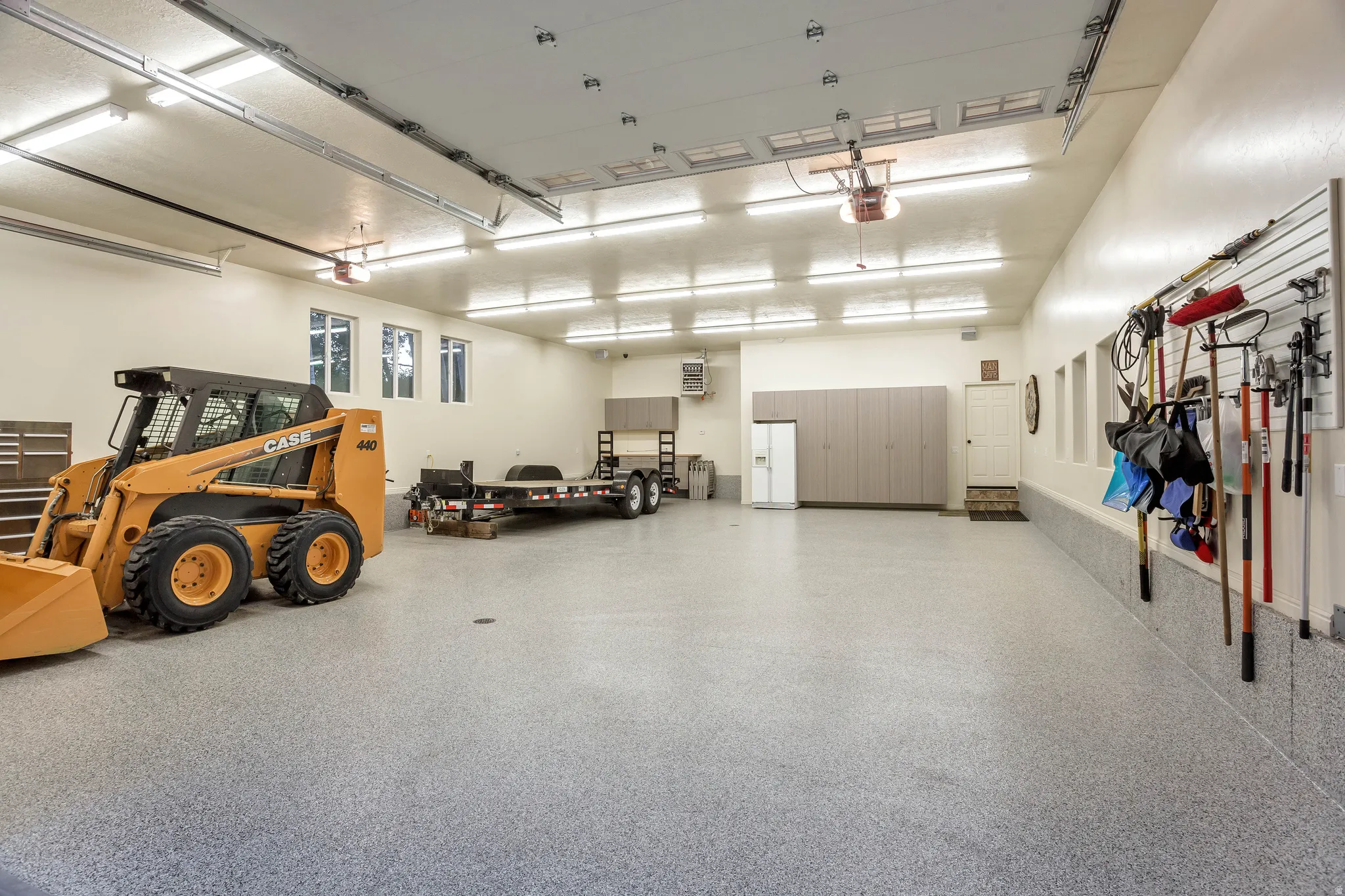 Spacious garage featuring an epoxy-coated concrete floor, ample overhead lighting, multiple windows, extensive built-in cabinetry, and slatwall storage systems
