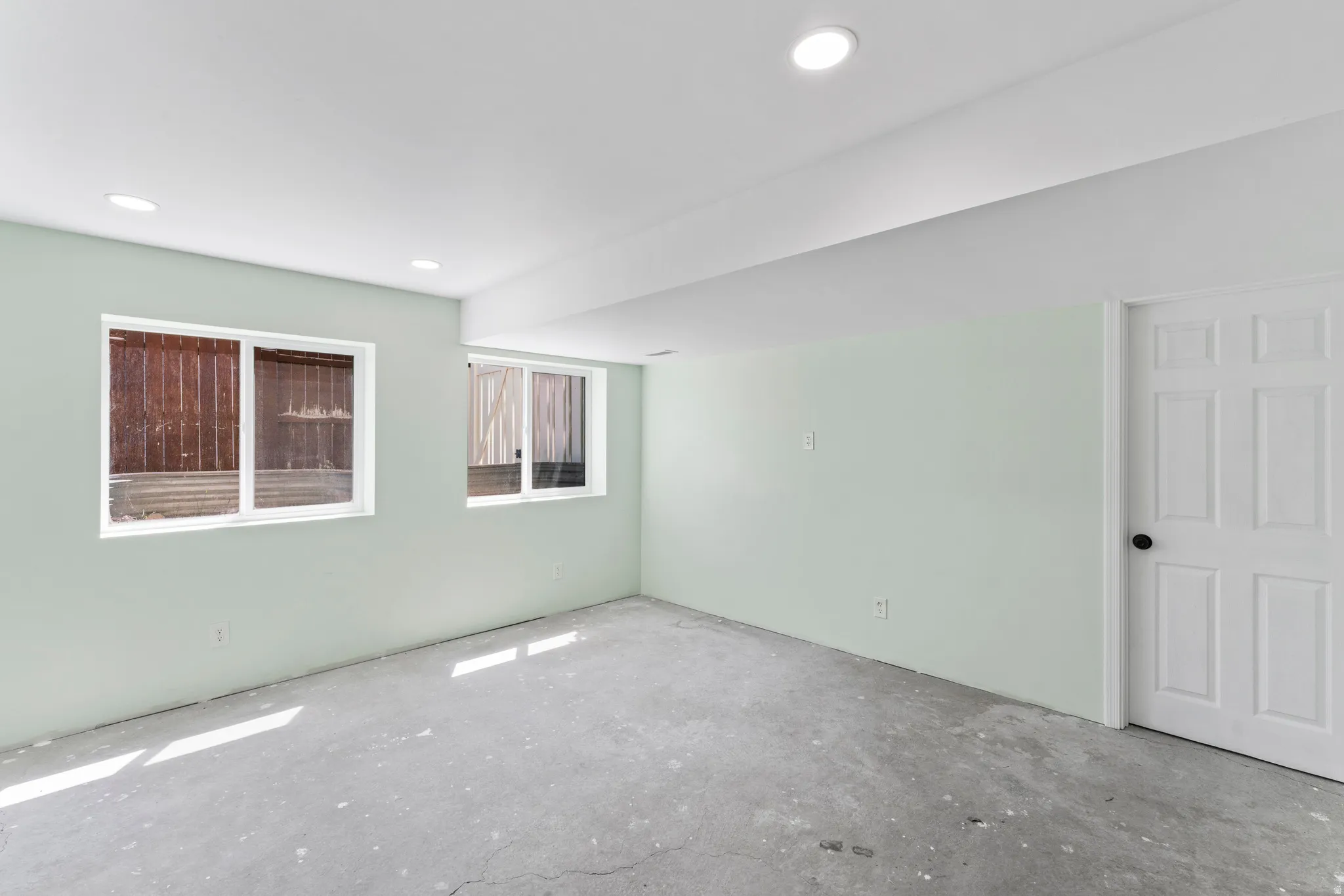 Spacious room featuring light green walls, two white-framed windows, recessed ceiling lighting, a white paneled door with a black doorknob, and concrete flooring