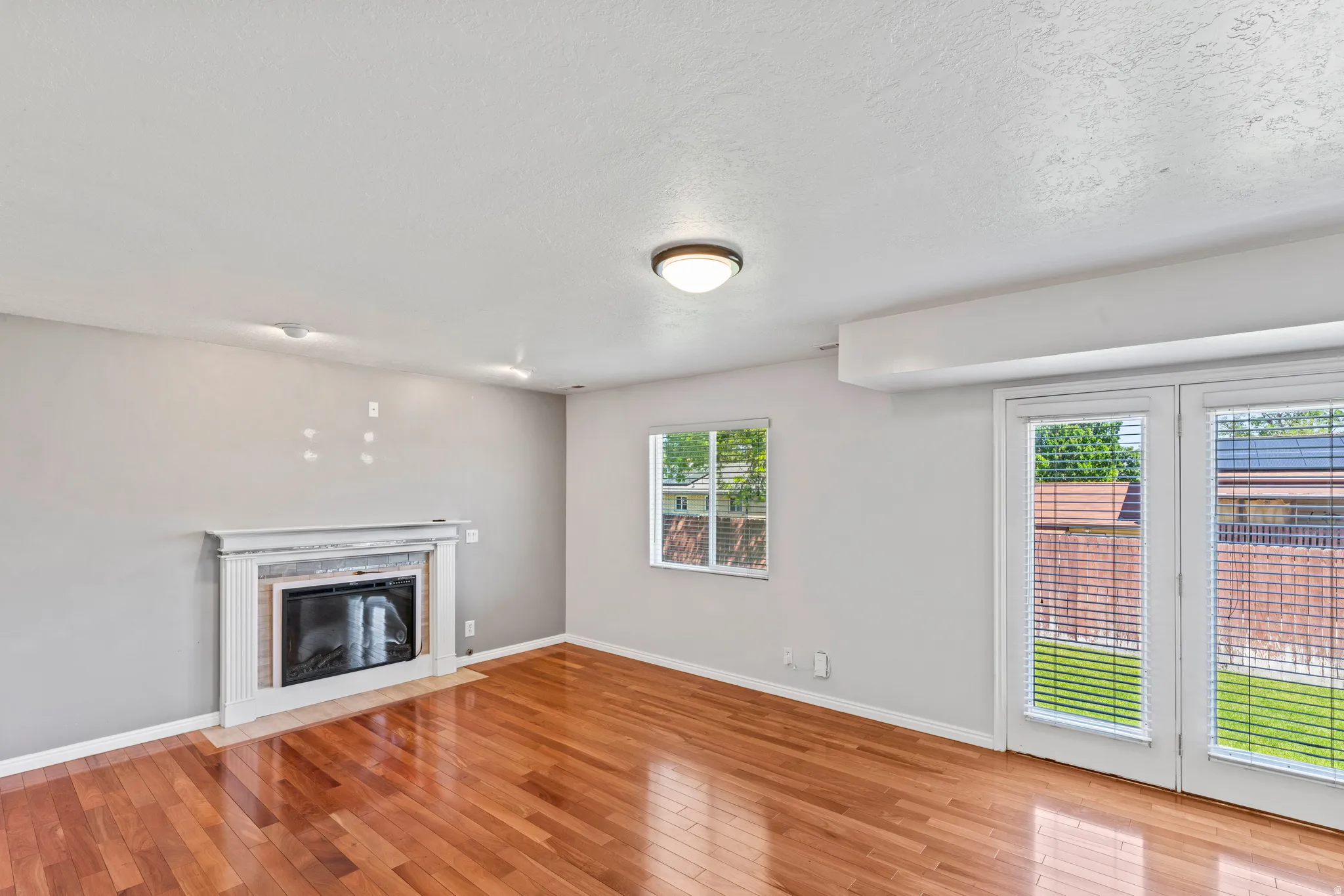 Living area featuring rich wood-finish flooring, a white mantel fireplace with tile surround, a window with blinds, and double glass panel doors providing direct outdoor access