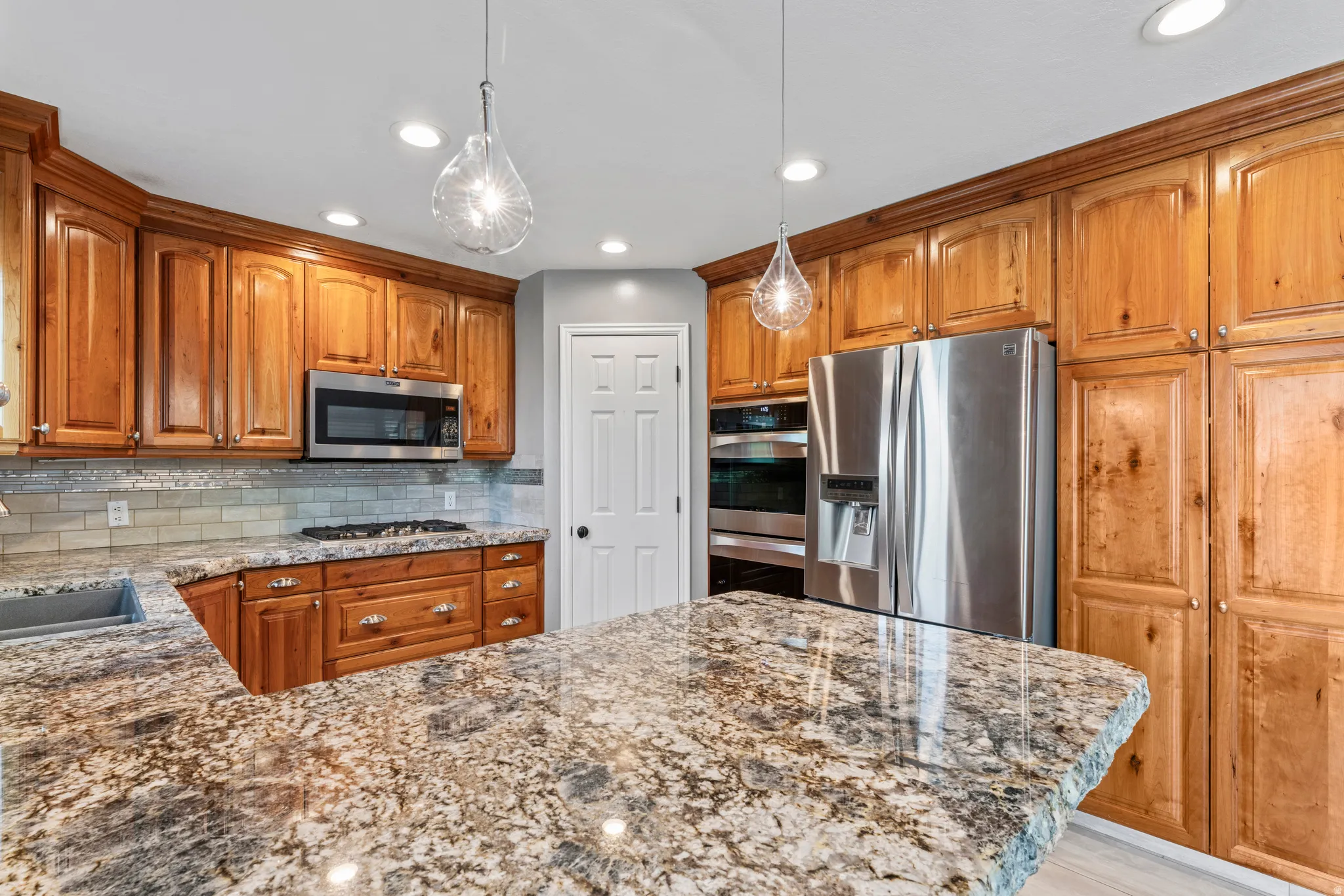 Spacious kitchen featuring light wood cabinetry, stone countertops, stainless steel appliances, a tile backsplash, and two pendant lights