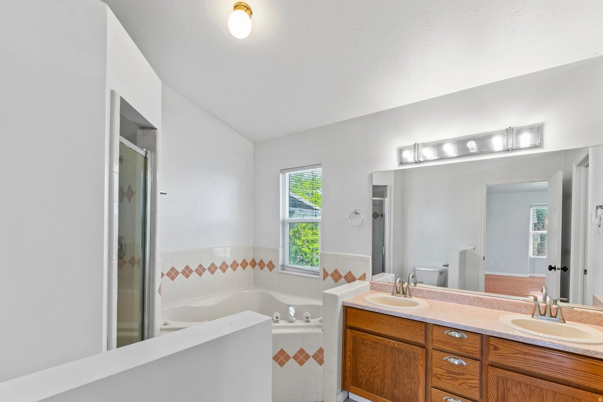 Bathroom featuring a dual vanity with oak-finish cabinetry and integrated sinks, a wide mirror with strip lighting, a corner jetted tub, and a glass-enclosed shower