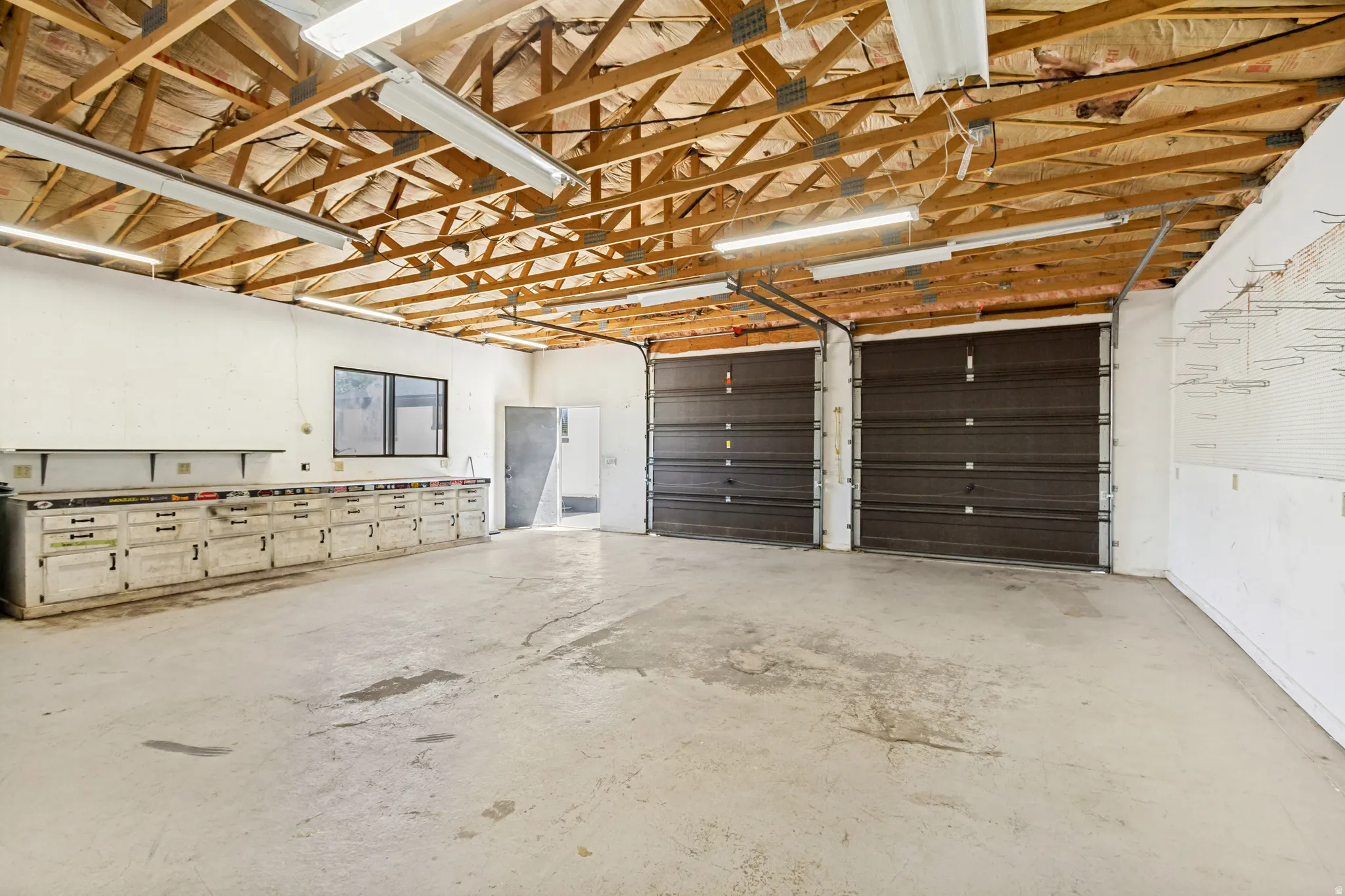 Spacious workshop featuring exposed wood beam ceilings, two large roll-up doors, extensive built-in cabinetry, dual-pane windows, and concrete flooring